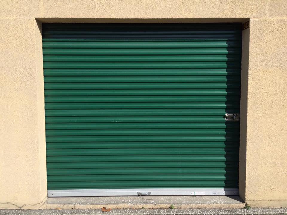 Green metal roll-up door against a beige wall, secured with a padlock, illustrating storage or commercial space access.