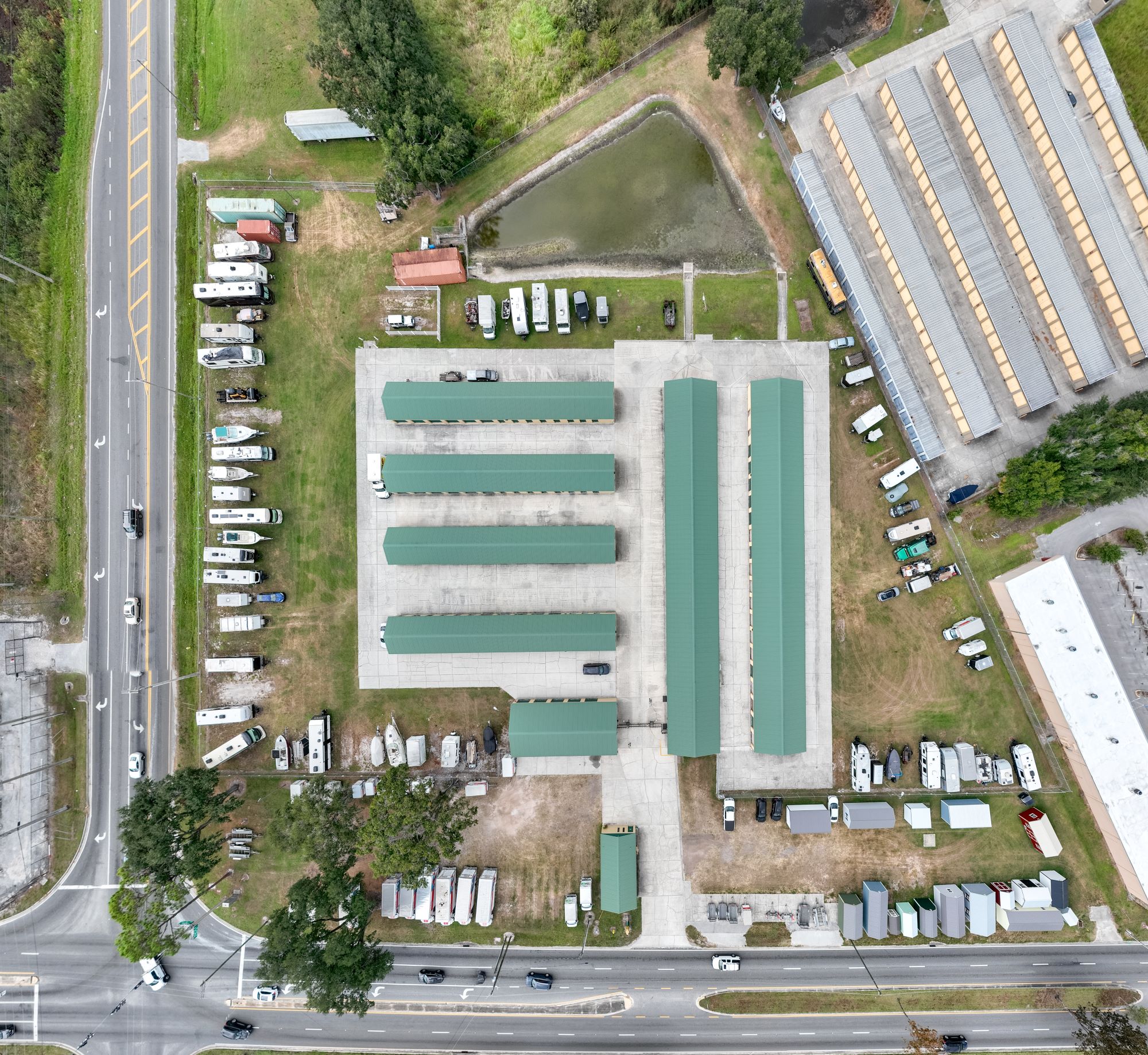 Aerial view of a storage facility featuring multiple covered parking areas for RVs and boats, surrounded by open land and a small pond.