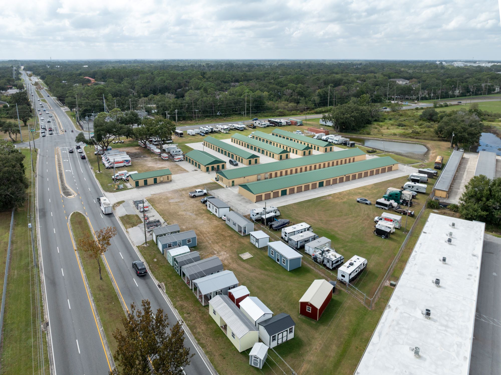 Aerial view of a storage facility with multiple units and RVs parked alongside a busy road, showcasing its layout and surrounding greenery.