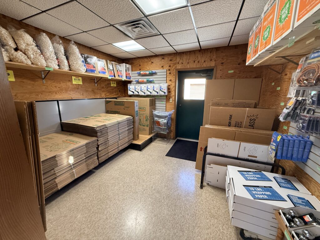 Packing supplies store interior showing stacked cardboard boxes, wrapping paper, and packing materials, with a door leading outside.